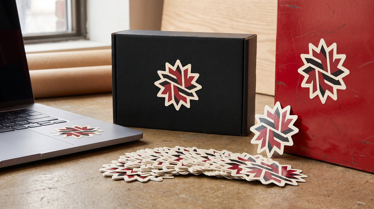 The same leaf-shaped die-cut sticker applied to three surfaces — a black mailer box, a red painted panel and the corner of a laptop — with a loose fanned pile of the same sticker on the desk in front